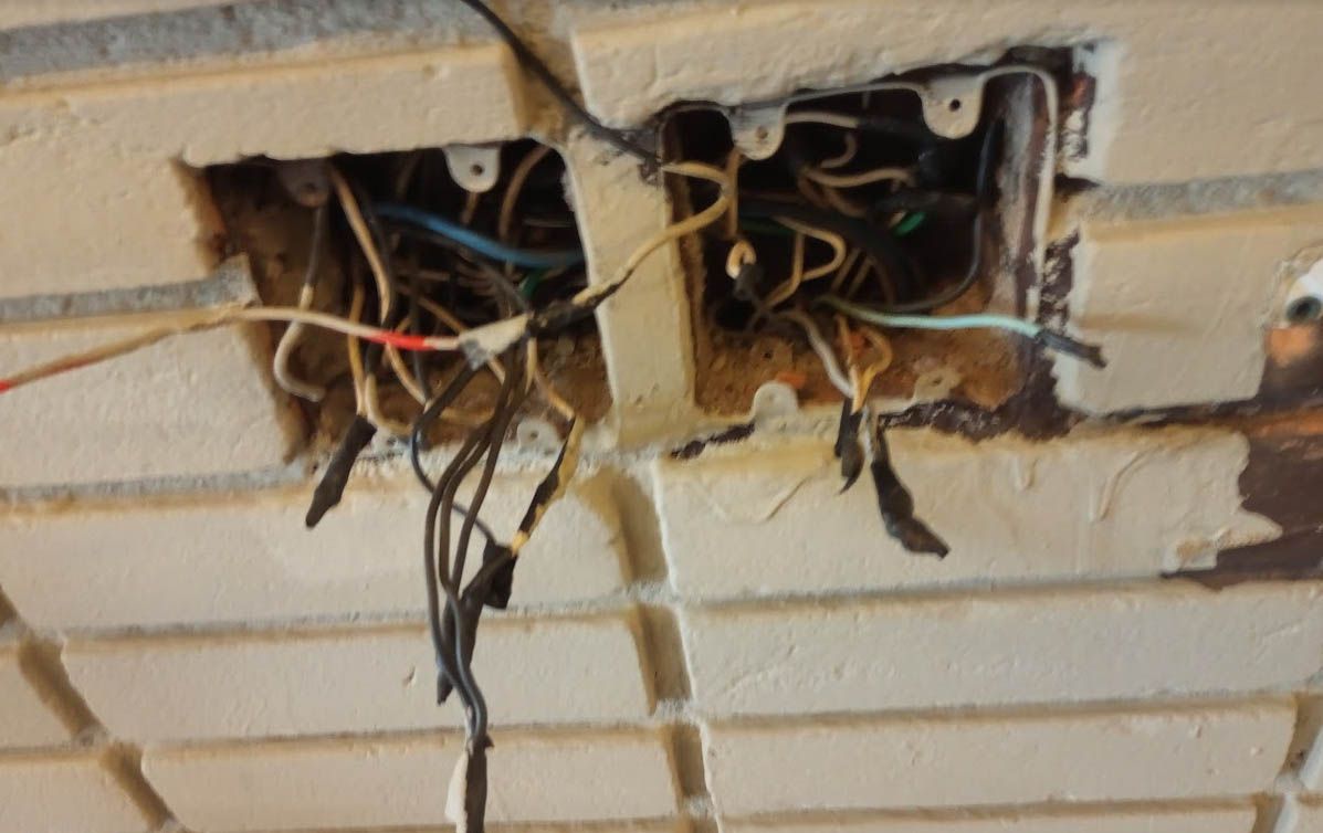 Instalação antiga dentro de parede de tijolos, com fios expostos, emendas feitas com fita isolante e caixas de passagem deterioradas